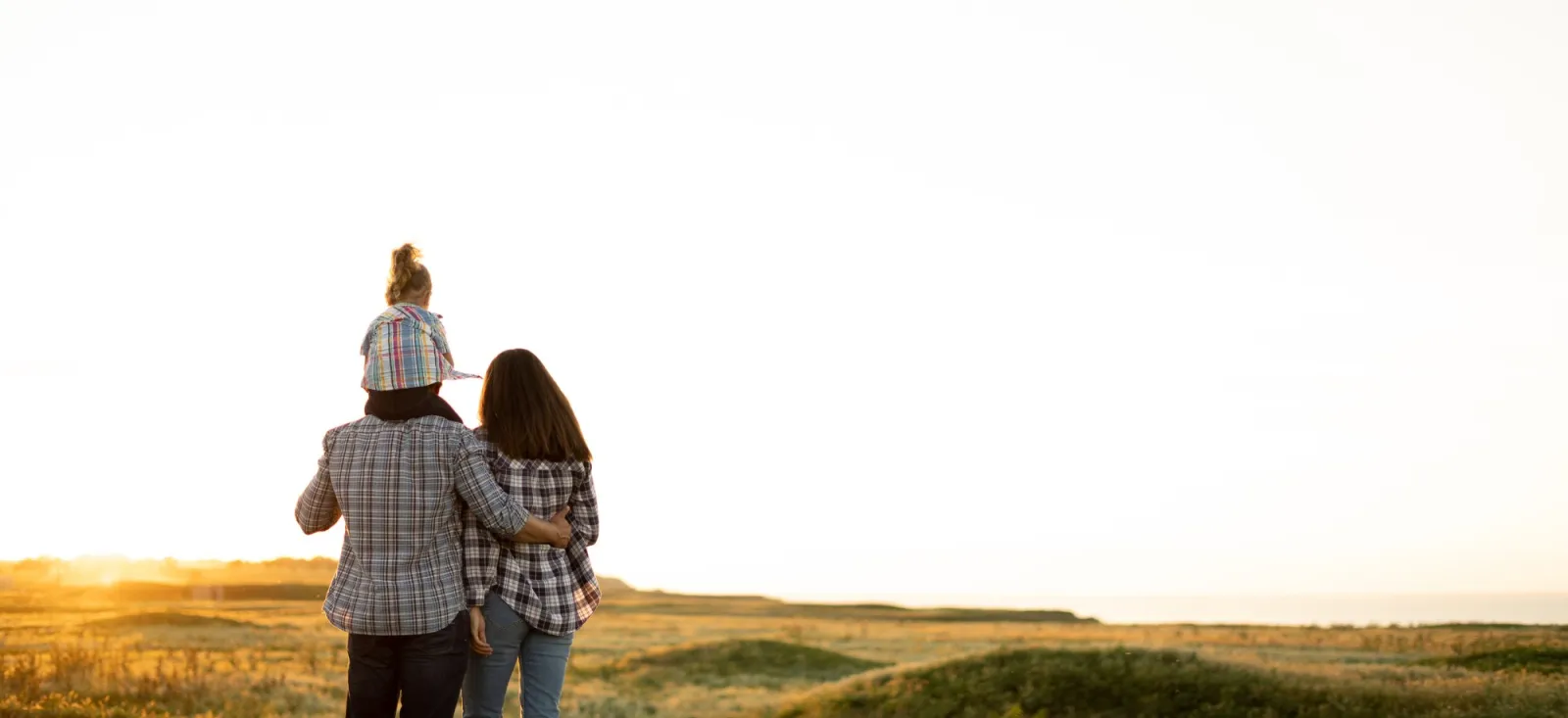 Family with a young child on a parent's shoulders watching the sunset over open fields